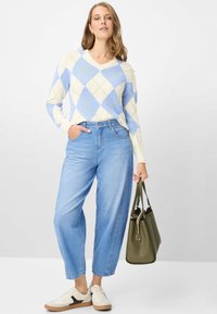 V-hals trui in blauw, crème en lichtgroen argylepatroon, gecombineerd met lichtblauwe wijde jeans en witte sneakers. Houdt een olijfgroene handtas vast.