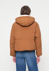 Geacă puffer maro cu un design scurt și glugă, având o interior moale de culoare albă și textură quilted. Asortată cu blugi albaștri.