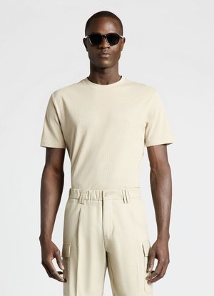 Homme portant des lunettes de soleil noires, une chemise beige à manches courtes et un pantalon cargo beige, debout devant un fond clair uni.