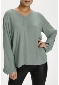 Blusa lunga verde chiaro, maniche lunghe in tessuto texture con scollatura a V, caratterizzata da un bottone decorativo e maniche leggermente svasate. Pantaloni neri indossati sotto.