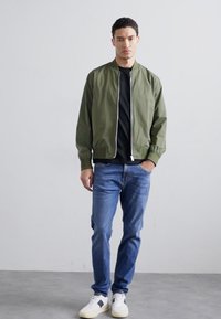 Vihreä pommijakku vetoketjulla edessä, musta T-paita alla, siniset farkut ja valkoiset tennarit. Sujuva kangas, rento malli, ribbaatut hihansuut.