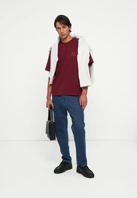 Camiseta de algodón burdeos con logo, combinada con jeans de mezclilla azules y zapatillas negras. Una chaqueta ligera está colgada sobre los hombros del modelo.