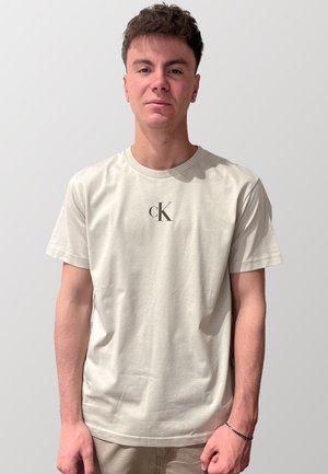 T-shirt in cotone beige chiaro con scollatura rotonda e maniche corte, caratterizzato da un logo "CK" nero stampato sul petto. Texture morbida.