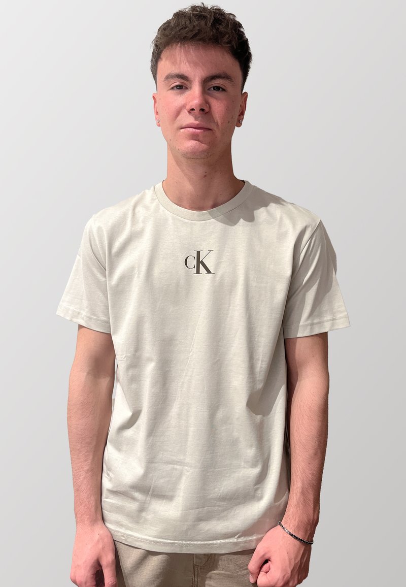 T-shirt in cotone beige chiaro con scollatura rotonda e maniche corte, caratterizzato da un logo "CK" nero stampato sul petto. Texture morbida.