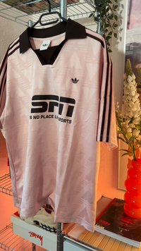 Φωτεινό ροζ αθλητικό μπλουζάκι Adidas με μαύρο γιακά και ρίγες κρεμασμένο σε μεταλλική κρεμάστρα κοντά σε λουλούδια και περιοδικά σε ένα ράφι.