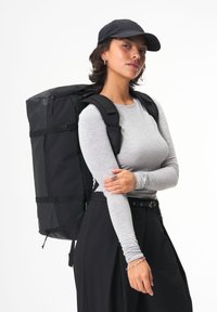 Schwarzer Rucksack aus langlebigem Material, mit verstellbaren Trägern, einem Reißverschluss oben und einer externen Tasche. Das Modell trägt ein graues Langarmhemd.