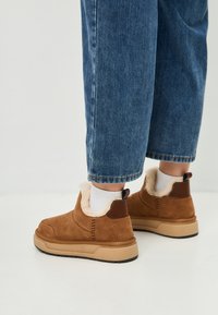 Piedi che indossano stivali alla caviglia in suede beige con fodera in pelliccia artificiale beige e calze bianche, abbinati a jeans in denim blu su uno sfondo semplice.