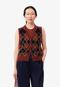 Gilet marrone senza maniche con un motivo a rombi neri e gialli, abbottonato sul davanti, indossato sopra una maglietta marrone coordinata. Pantaloni scuri, logo visibile.