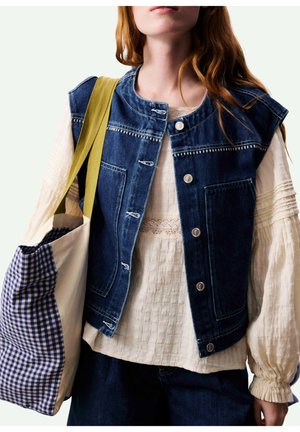 Femme portant une blouse blanche texturée et un gilet en denim bleu, portant un sac fourre-tout crème avec une doublure vichy bleue et des sangles jaunes.