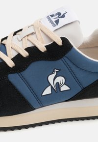 Zapatilla con un upper de ante negro, paneles de textil azul, cordones color crema y un logo blanco en la lengüeta. Suela de goma con detalles de tracción.