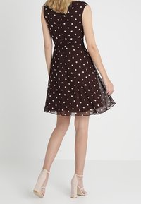 Ruskea polkadot-printtimekko, jossa liehukehelma, hihaton muotoilu ja vyötäröllä kapea laskos. Täydennetty vaaleanbeigeillä, paksukorkoisilla sandaaleilla.