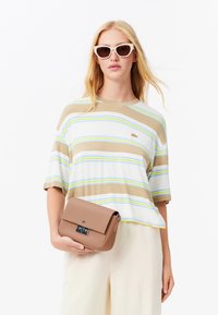 Camicia a maniche corte a righe in beige, bianco e blu; abbinata a una borsa a tracolla marrone chiaro con una chiusura metallica. Il modello indossa occhiali da sole a occhio di gatto.