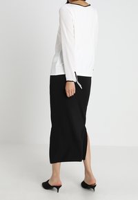 Schwarze Rock mit seitlichem Schlitz, kombiniert mit einem weißen Langarmshirt mit schwarzem Besatz und Schleifen an den Ärmeln. Glatte Textur, minimalistisches Design.