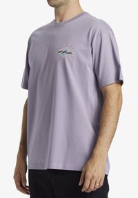 Camiseta de algodón lavanda con manga corta, que presenta un diseño gráfico circular con acentos azules y marrones en el pecho.