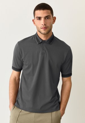 Bărbat cu păr scurt și închis la culoare, purtând un tricou polo gri cărbune cu model negru pe guler și mâneci, stând cu mâinile în buzunare.