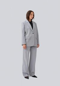 Completo grigio chiaro su misura con giacca a doppiopetto e pantaloni a gamba larga. Texture liscia, design formale, abbinato a una camicia nera e scarpe a punta.