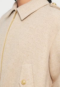 Casaco bege com fecho de correr, feito de tecido texturizado, com gola, fecho de correr dourado e um bolso no peito com um botão dourado de destaque.