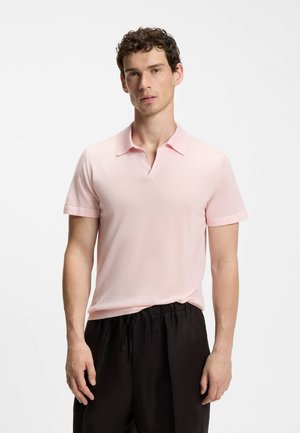 Uomo con capelli corti e ricci indossa una polo rosa chiaro a maniche corte e pantaloni neri larghi, in piedi contro uno sfondo bianco semplice.