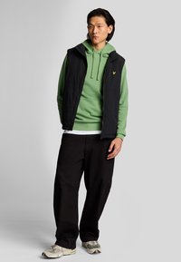 Gilet nero senza maniche sopra un hoodie verde, abbinato a pantaloni neri larghi e sneakers beige. Il gilet ha una chiusura con zip e tasche.