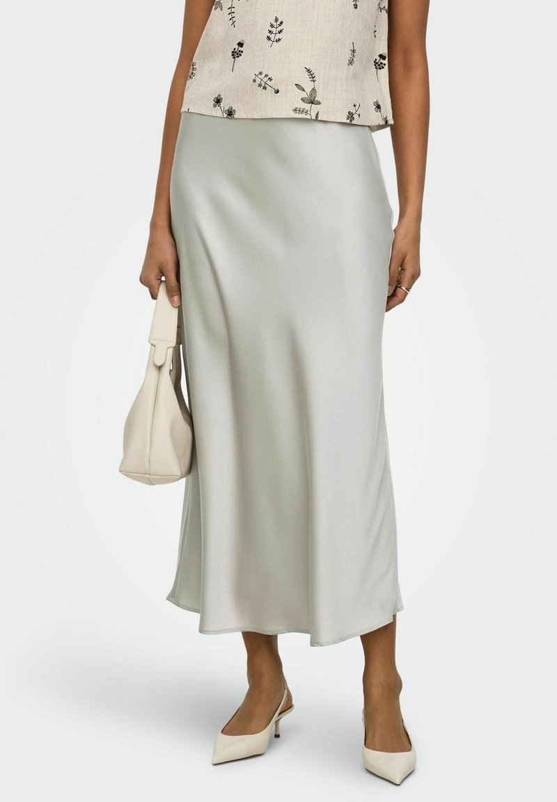 Femeie purtând o fustă midi din satin gri deschis, top bej floral fără mâneci, pantofi cu bretea albă și toc, ținând o geantă crem pe lângă corp.