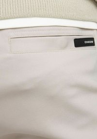 Beige, soepel stoffen broek met een achterzak met een zwart label waarop "CHASIN'" staat. Eenvoudig ontwerp, zonder zichtbare patronen of texturen.