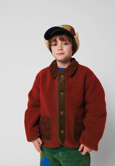 Giacca in pile rossa con accenti in corduroy marrone, grandi bottoni frontali e due tasche. Il modello indossa un cappello multicolore e pantaloni in corduroy verdi.