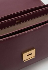 Borsa in pelle viola con cuciture bianche, apertura superiore che mostra l'interno in tessuto e una chiusura rettangolare dorata con la scritta MNG.