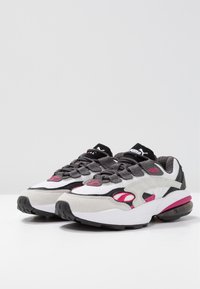 Sneakers sportive con una toma superiore in rete bianca, accenti in suede grigi, dettagli rosa e una suola in gomma nera con un'unità d'aria visibile.