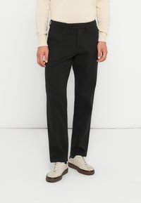 Sorte bukser med lige pasform, lavet af et glat stof. Har en knaplukning og bæltestropper, parret med beige sneakers.