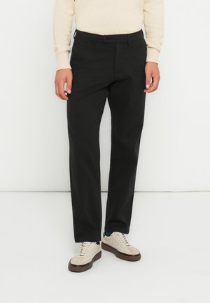 Pantalon noir à coupe droite, fabriqué dans un tissu lisse. Dispose d'une fermeture à bouton et de passants de ceinture, associé à des baskets beiges.