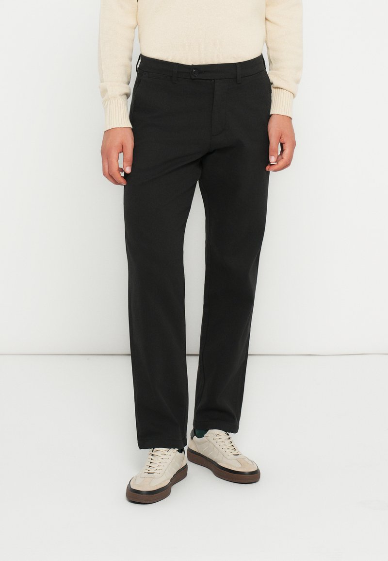 Sorte bukser med lige pasform, lavet af et glat stof. Har en knaplukning og bæltestropper, parret med beige sneakers.