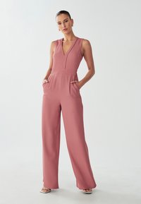 Roze mouwloze jumpsuit met een diepe V-hals, taps toelopende taille en een ontwerp met wijde pijpen. Kenmerkt zich door zijzakken en een gladde, zachte textuur.