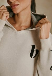 Beige sweater med hætte, der har et sort broderet bogstav "b". Stoffet ser blødt ud med et struktureret indre foer.