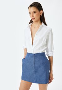 Chemise blanche à boutons avec des manches retroussées, associée à une mini-jupe bleue texturée présentant une coupe ajustée et des poches latérales.