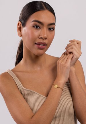 Goldene Kettenarmband mit einem runden Anhänger, der eine texturierte Oberfläche aufweist, am Handgelenk getragen. Das Modell hat glattes, dunkles Haar und trägt ein Top in neutralen Tönen.
