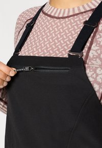 Melni pārvalki ar regulējamām siksnām, ar iešūtu kabatu uz krūšu. Cilvēks nēsā apakšā rakstainu rozā blūzi.