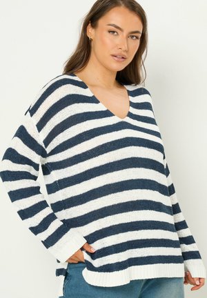 Kvinde iført marineblå og hvide brede stribede V-hals trøje med ribstrikkede manchetter og kant, kombineret med blå jeans, foran en lys baggrund.