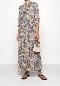 Robe maxi florale avec des manches trois quarts, ornée d'un mélange de fleurs roses, bleues et vertes. Assortie à un petit sac à main beige clair et des sandales plates.