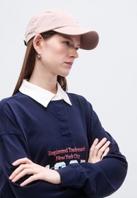 Ροζ ανοιχτό ροζ καπέλο με καμπυλωτό γείσο, φορεμένο με μπλε πουκάμισο πόλο με γιακά και κόκκινη κεντητική. Μοντέλο που εμφανίζει διασταυρωμένα τα χέρια του πάνω σε ένα ουδέτερο φόντο.