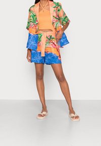 Femme portant un blazer à imprimé tropical vibrant et un short assorti avec un crop top halter orange et des sandales à enfiler beiges.
