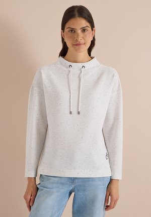 Femme aux cheveux foncés portant un pull blanc à manches longues tacheté avec des cordons et un jean bleu clair, debout devant un fond beige.