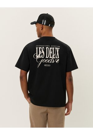 Camiseta negra de manga corta con texto blanco en la parte de atrás que dice "LES DEUX GOODS" con acentos. Tela suave y corte relajado.