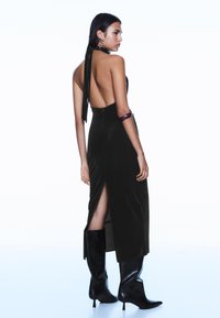 Czarna aksamitna sukienka na szyję z odkrytymi plecami, rozcięciem z boku i czarnymi wysokimi butami na obcasie. Modelka nosi fioletową bransoletkę i kolczyki.