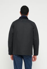 Casaco isolado preto com gola em bombazina azul-marinho, apresentando um logótipo nas costas, mangas compridas e corte descontraído, combinado com jeans azuis.