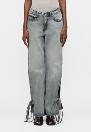 Άτομο που φοράει γκρι τζιν με φαρδιά μπατζάκια και πλευρικά σκισίματα δεμένα στα τελειώματα, συνδυασμένα με σκούρα παπούτσια με τετράγωνη μύτη και ανοιχτόχρωμο αμάνικο τοπ.