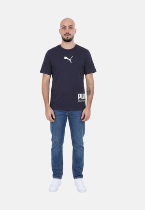 Uomo con barba e tatuaggio che indossa una maglietta Puma blu navy, jeans blu e sneakers bianche, in piedi davanti a uno sfondo chiaro e uniforme.