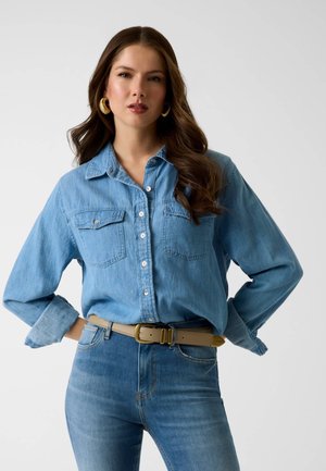 Donna con capelli castani lunghi che indossa una camicia di denim blu, jeans blu, cintura beige e grandi orecchini a cerchio dorati, in piedi con le mani sui fianchi.