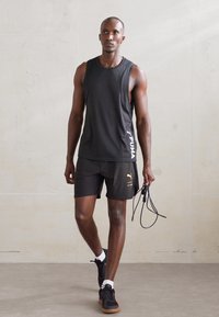 Zwart mouwloos sporttop en shorts, beide met Puma-logo's in wit en goud. Model houdt een springtouw vast; zwarte sneakers met een rubberen zool.