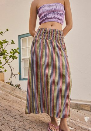 Frau trägt ein pastell-lila gerafftes Bandeau-Top und einen hoch taillierten, bunt gestreiften Midi-Rock und steht auf einer gepflasterten Straße.