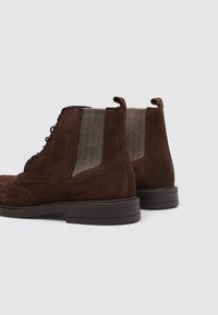Las botas de tobillo de ante marrón cuentan con un diseño de brogue, paneles laterales de textil de espiga y una suela de goma resistente. Tacón reforzado con una lengüeta.